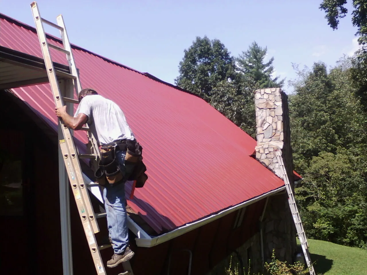 Roofer on a ladder beside a red metal roof and stone chimney — Soffit & Fascia Repair by Roofing Brothers in Tinley Park